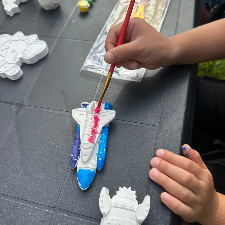 Mains d'enfant peignant une figurine de navette spatiale en plâtre, activité du Pack Anniversaire RIIN Art